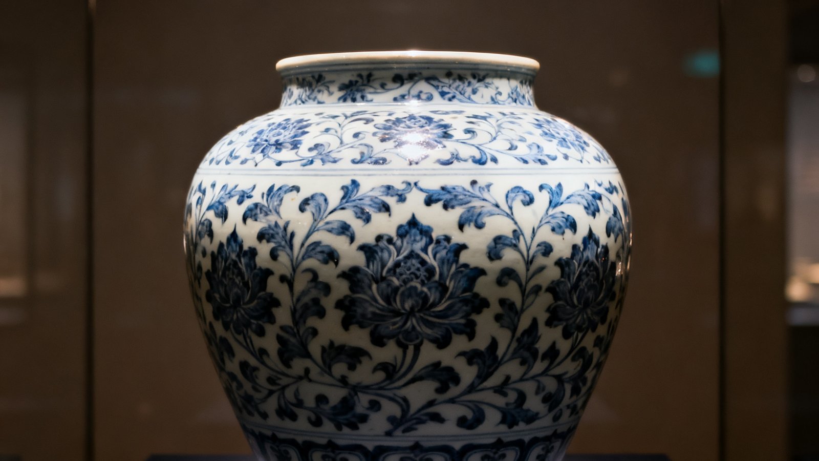 一件明代宣德年间的青花缠枝莲纹大罐置于深色展台上，灯光从上方柔和洒落，凸显出瓷釉的温润光泽与精细的笔触细节。